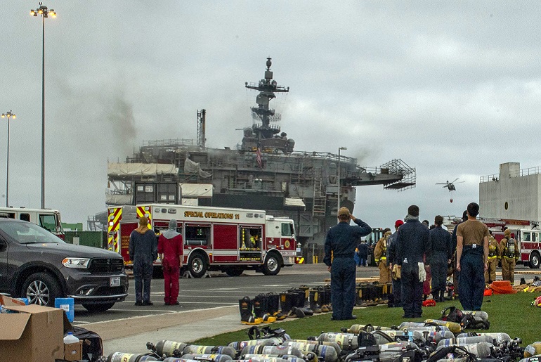 Membros da guarnição do navio perfilam-se em sinal de respeito durante o hastear da bandeira, ao mesmo tempo que decorrem as operações de combate ao incêndio (imagem US Navy)